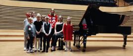 Na estradzie sali koncertowej nastolatka siedzi na krześle obok fortepianu stojącego po prawej stronie, wokół niej stoi pięć dziewczynek i trzech chłopców