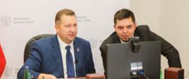Minister Czarnek i mężczyzna w garniturze siedzą przed monitorem i rozmawiają.