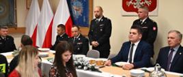 na zdjęciu widoczny Komendant Miejski wraz z Prezesem Oddziału Powiatowego ZOSP, obok zaproszeni goście, w tle godło Rzeczypospolitej Polskiej oraz poczet sztandarowy
z flagami
