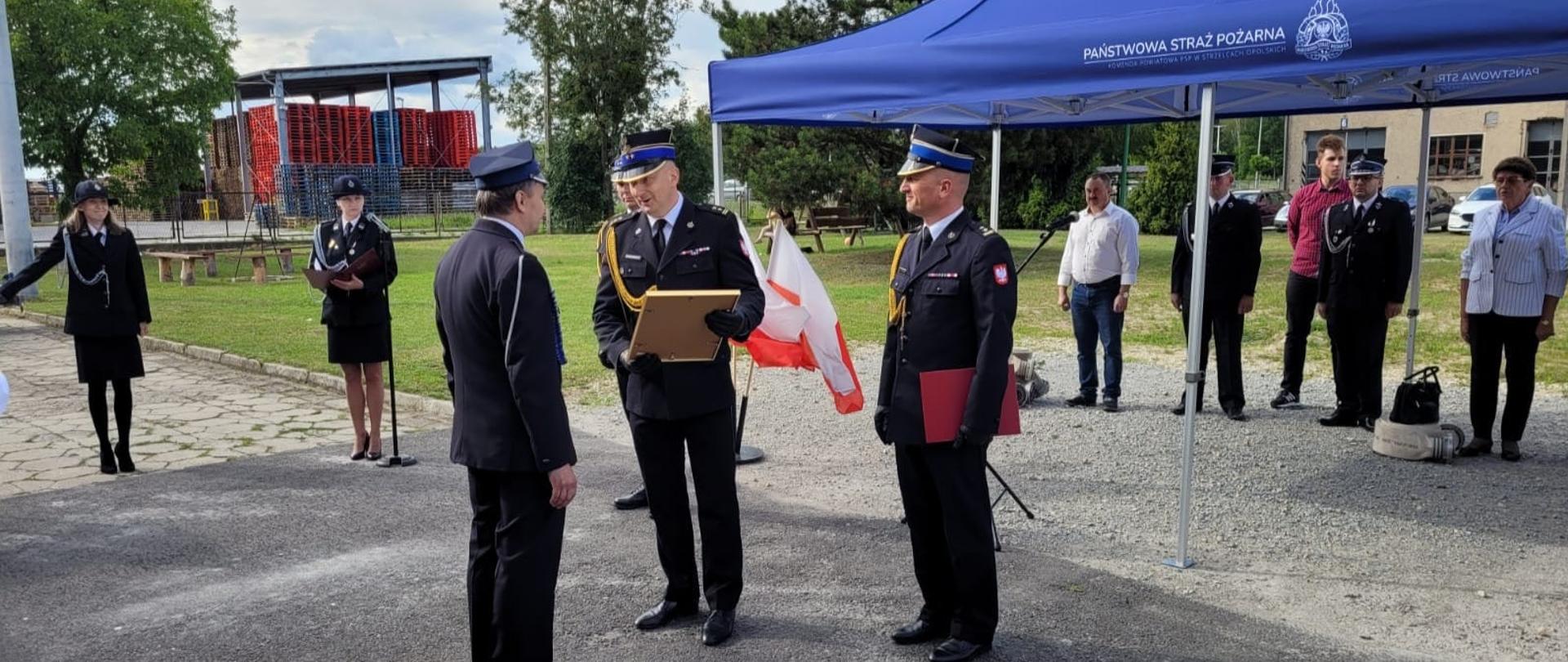 Bryg. Dariusz Gieroń p.o. Zastępca Opolskiego Komendanta Wojewódzkiego PSP w Opolu w towarzystwie bryg. Pawła Poliwody Komendanta Powiatowego PSP w Strzelcach Opolskich, wręcza decyzję o włączeniu jednostki OSP Strzelce Opolskie do krajowego systemu ratowniczo-gaśniczego Prezesowi OSP.
Komendanci, druhny i druh Prezes w mundurach galowych. Pozostali goście zgromadzeni pod namiotem KP PSP w Strzelcach Opolskich.
