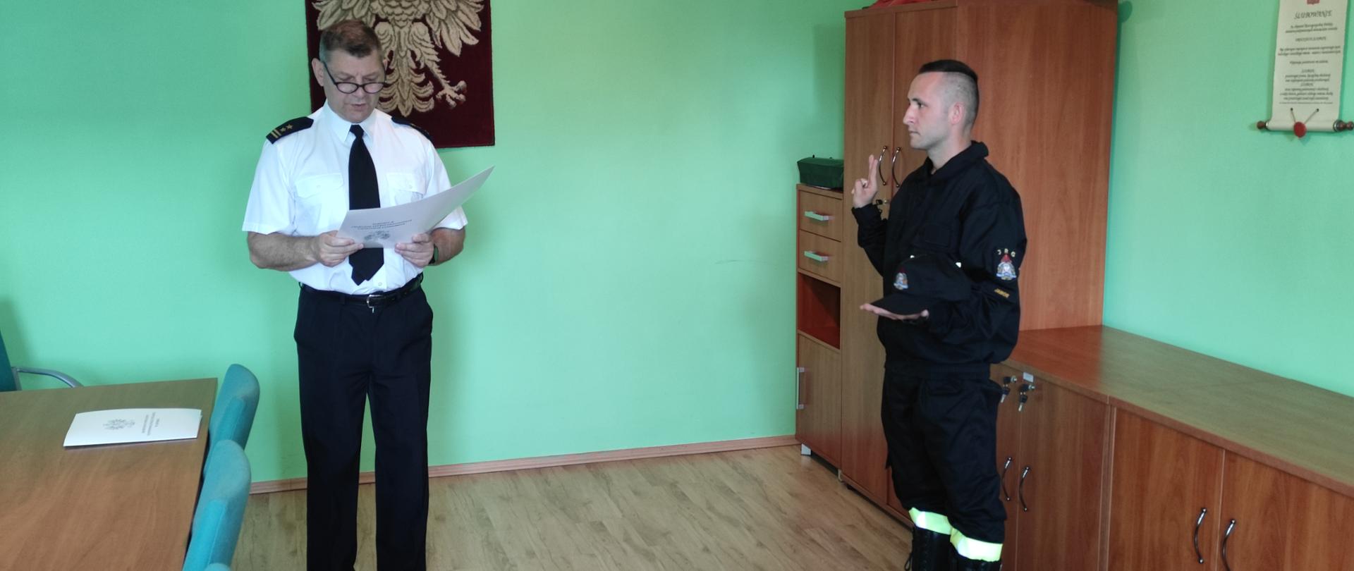 Obraz przedstawia strażaka podczas uroczystego ślubowania. Komendant odczytuje słowa roty ślubowania. Strażacy w sali narad