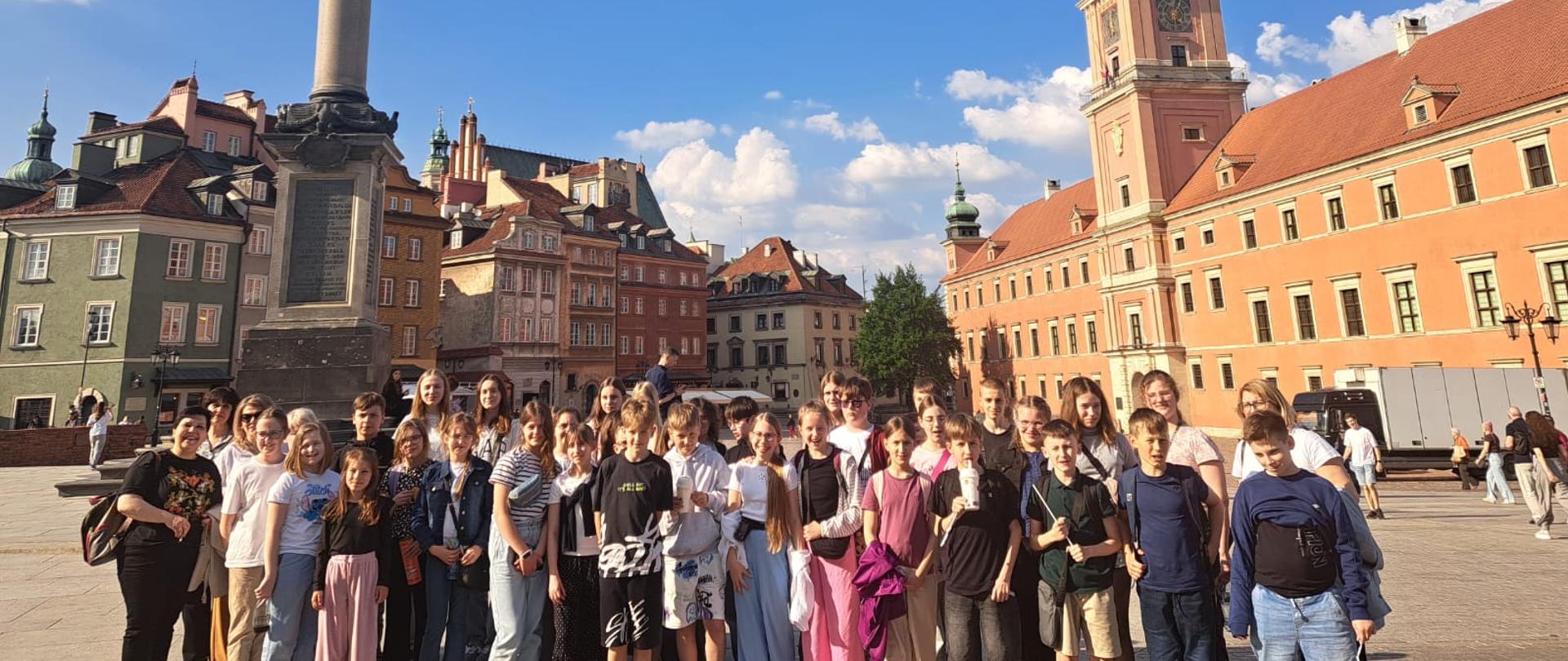 Grupa dzieci i młodzieży wraz z opiekunką pozuje do wspólnego zdjęcia na dużym placu miejskim. W tle widoczne są zabytkowe kamienice Warszawy, Zamek Królewski oraz Kolumna Zygmunta Uczestnicy stoją w kilku rzędach i patrzą w stronę aparatu. Zdjęcie wykonano w słoneczny dzień przy bezchmurnym, błękitnym niebie