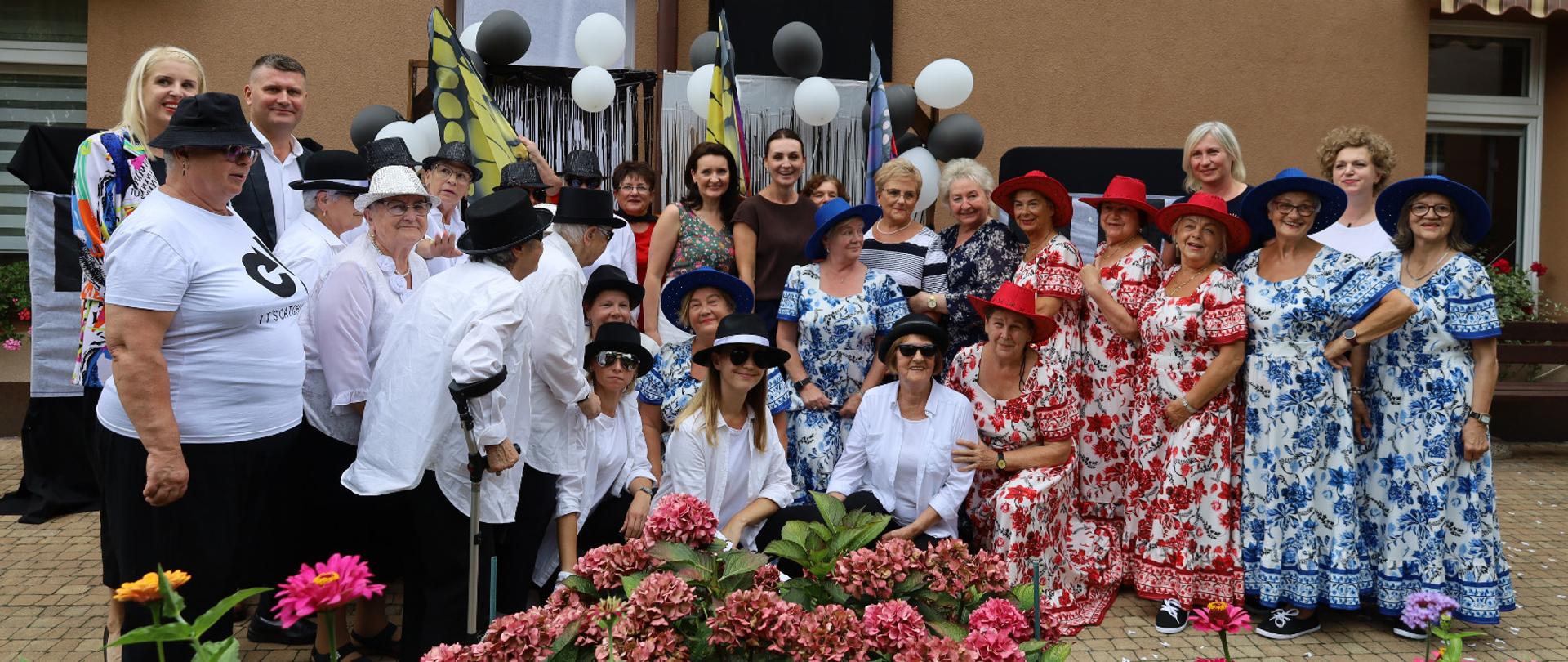 grupa osób pozuje do zdjęcia, kobiety maja podobne wzorzyste suknie i kapelusze, pośrodku stoi minister i wojewoda