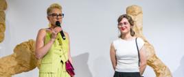 Dwie kobiety w przestrzeni wystawowej. Za nimi duże obiekty rzeźbiarskie z papieru. Kobieta po lewej mówi do mikrofonu