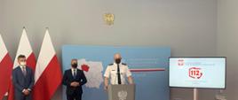 Zdjęcie przedstawia 3 mężczyzn znajdujących się w pomieszczeniu, w którym odbywa się konferencja prasowa. Znajdują się tam również 3 flagi Polski, mównica, baner, telewizor na którym widać odtwarzaną prezentację oraz kamera