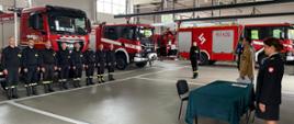 Strażacy na garażu KP PSP w Ciechanowie. Po lewej stronie strażacy w ubraniach koszarowych na tle samochodów gaśniczych, po prawej stronie ślubujący strażak, sztandar komendy oraz inni fukcjonariusze