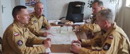 Zdjęcie przedstawia odprawę operacyjną w biurze komendanta, strażacy siedzą przy stole gdzie sa mapy.