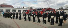 Zdjęcie przedstawia strażaków z Państwowej i Ochotniczej Straży Pożarnej w mundurach wyjściowych. Strażacy trzymają otrzymane promesy w rękach. W tle znajdują się samochody pożarnicze koloru czerwonego oraz magazyn