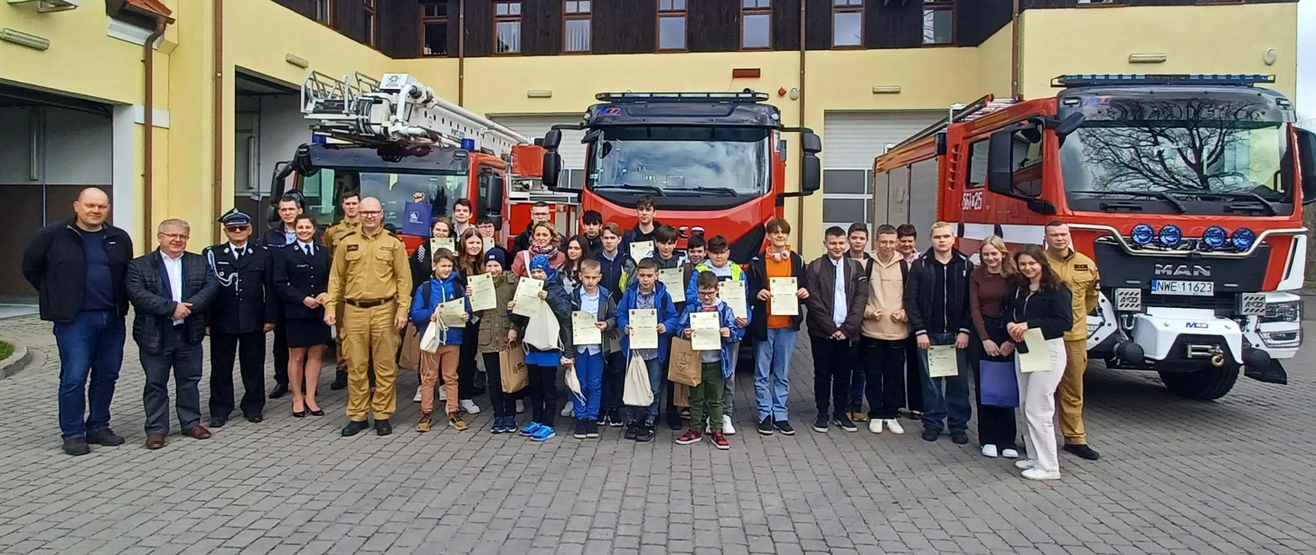 Uczestniczy konkursu wraz z opiekunami stoją na placu KP, za nimi ustawione są pojazdy pożarnicze i widoczny jest budynk KP. 