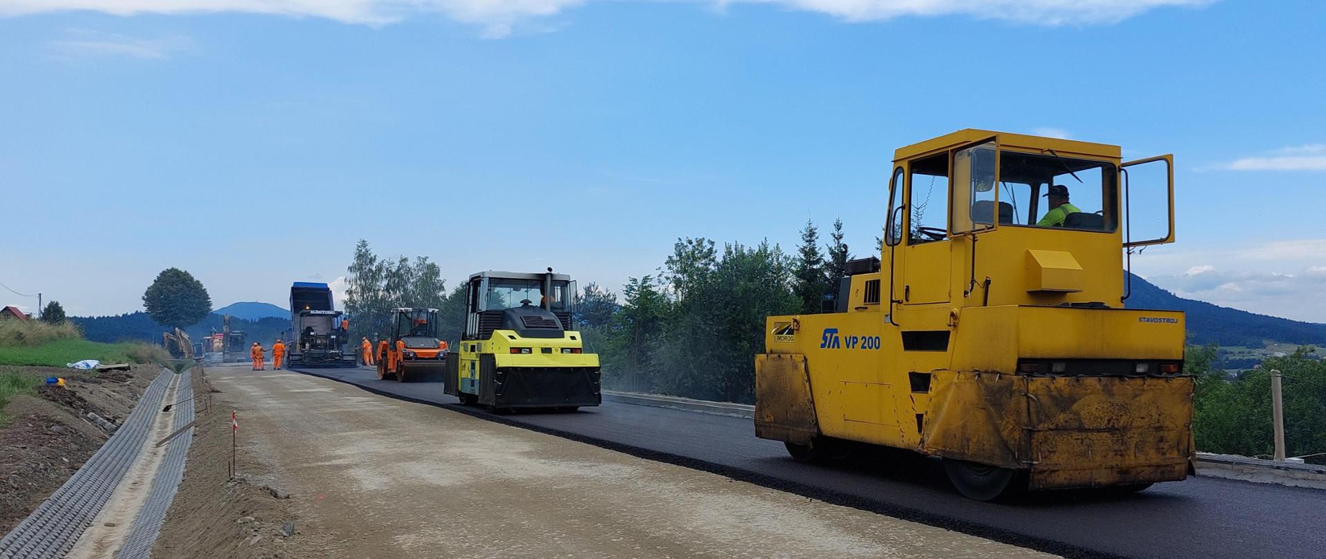 Na nowo wyłożonym asfalcie widoczne są trzy pracujące walce wyrównujące wylaną warstwę asfaltu. W tle można dostrzec kilku pracowników w ubraniach odblaskowych oraz kolejny pojazd wylewający asfalt na drogę. Niebo jest częściowo zachmurzone, a otoczenie wydaje się wiejskie, z zielenią po obu stronach drogi.