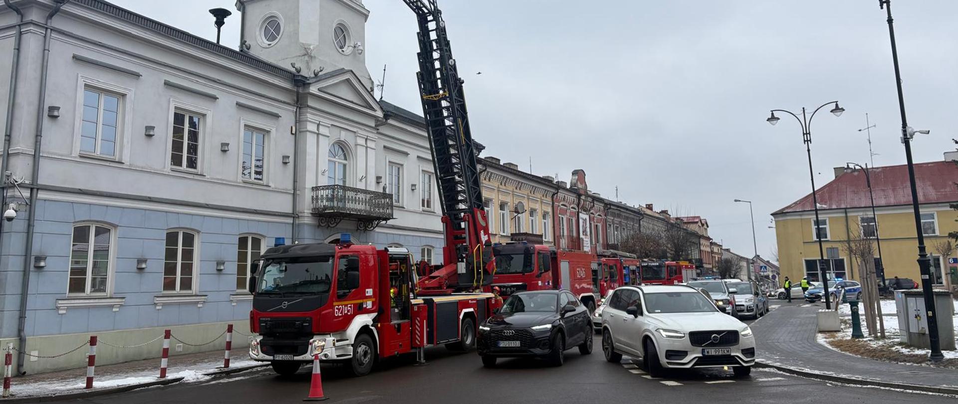 Czerwony wóz strażacki z wysuniętą drabiną mechaniczną stoi na ulicy przed zabytkowym budynkiem z wieżą zegarową. Na podnośniku znajdują się strażacy pracujący przy elewacji, a obok przejeżdżają samochody i widać zabezpieczony fragment jezdni.