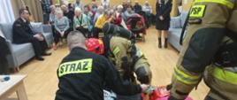 Pokaz udzielania pomocy osobie zaczadzonej wykonywany przez strażaków ubranych w aparaty tlenowe w tle publiczność zgromadzona na sali.