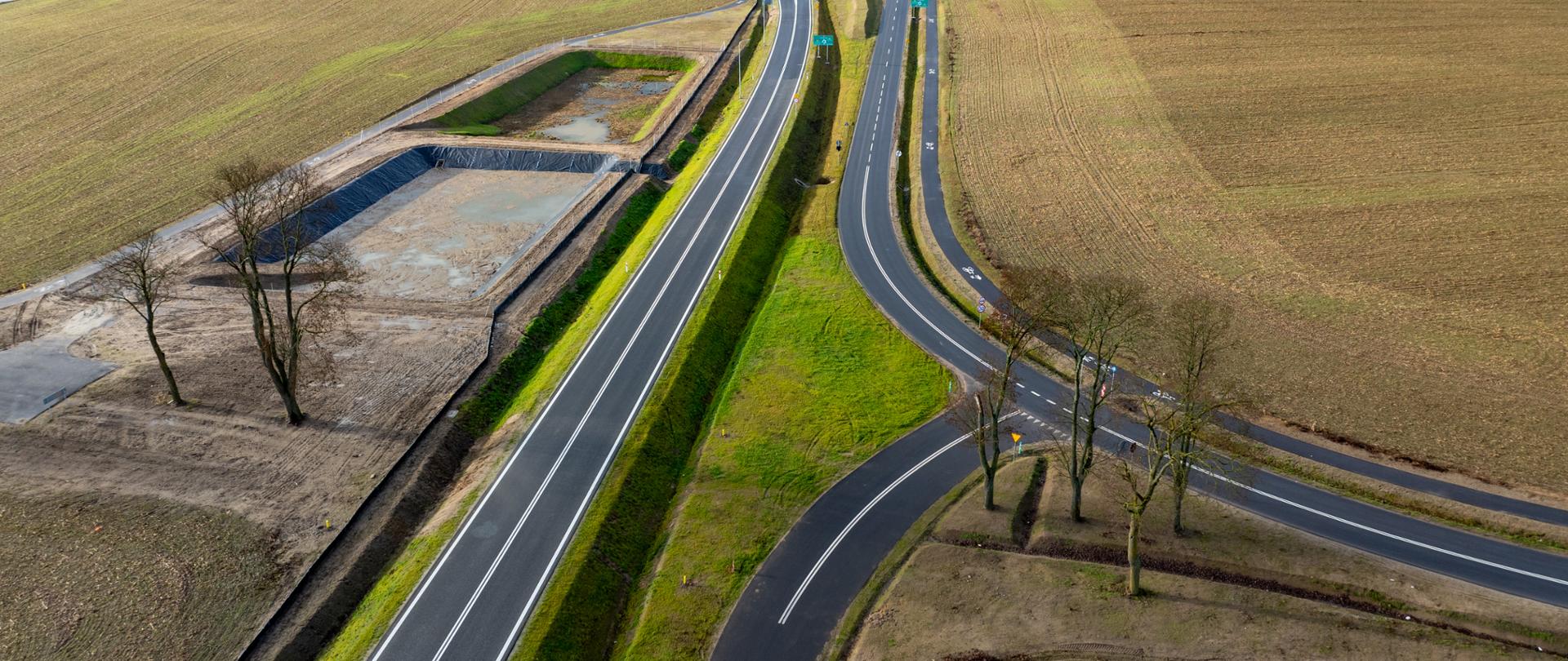 Panorama z lotu ptaka ukazująca świeżo wybudowaną obwodnicę biegnącą przez rozległe pola. Droga jest jednopasmowa, z wyraźnymi białymi liniami i łagodnymi zakrętami. Z prawej strony biegnie droga lokalna z poprowadzoną obok ścieżką rowerową. Po lewej stronie widoczny jest zbiornik retencyjny, a wzdłuż trasy rosną pojedyncze drzewa.