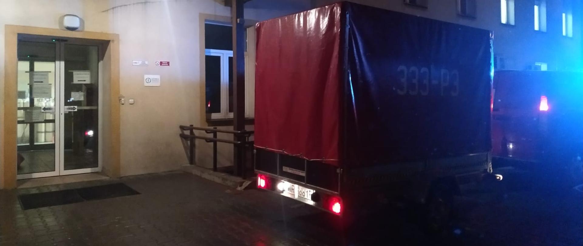 Zdjęcie przedstawia pojazd pożarniczy dostawczy typu furgon. Do pojazdu podpięta jest przyczepka. Pojazd jak i przyczepka są w kolorze czerwonym Pojazd posiada włączone niebieskie światła alarmowe. Wraz z przyczepką stoi na podjeździe przed wejściem głównym do szpitala w Pionkach. Zdjęcie wykonane z bliższej odległości w stosunku do poprzedniego oraz wykonane w porze nocnej. 