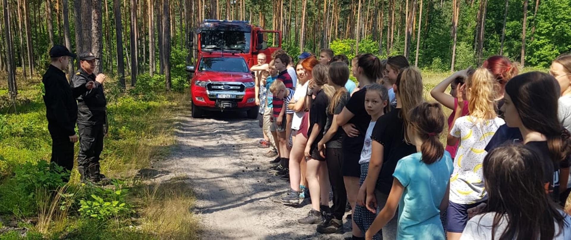 Zdjęcie przedstawia grupę dzieci i młodzieży na drodze leśnej podczas próbnej ewakuacji. W tle widać samochody straży pożarnej