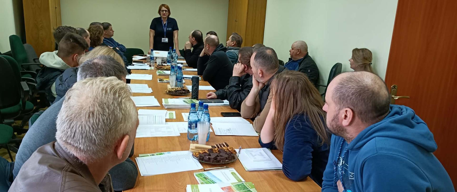 grupa osób siedzi naprzeciw siebie przy stole patrząc na stojąca w głębi zdjęcia kobietę