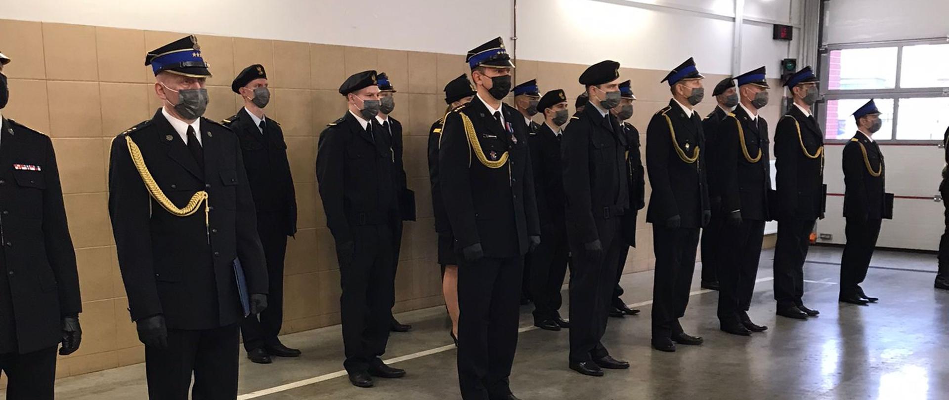 Część wyróżnionych strażaków stojąca w pododdziale w ubraniach wyjściowych. Na głowach czapki rogatywki, berety, Panie mają kapelusze, wszyscy maski na twarzach