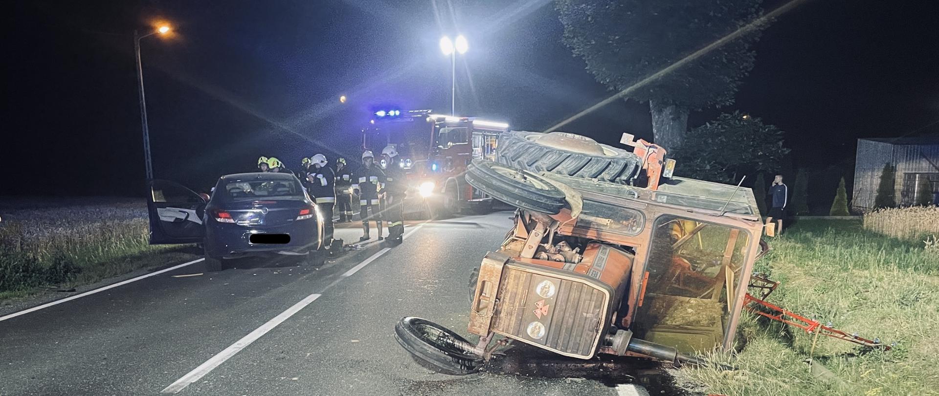 Na zdjęciu widoczny przewrócony ciągnik rolniczy oraz samochód osobowy po zderzanie w dali samochód pożarniczy oraz ratownicy