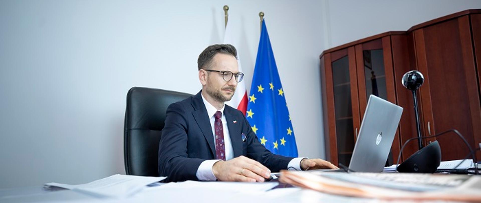 Wiceminister Waldemar Buda siedzi przy biurku przed laptopem. Z tyłu widoczne flagi UE i Polski.
