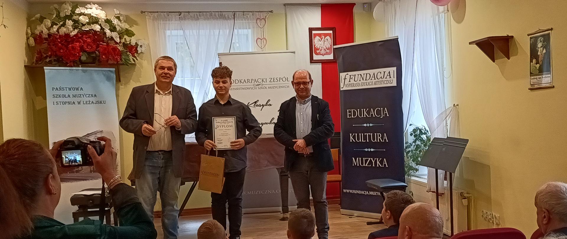 Na zdjęciu uczeń z organizatorami konkursu ,uczeń trzyma dyplom w ręku , na dole zdjęcia widać widownię.