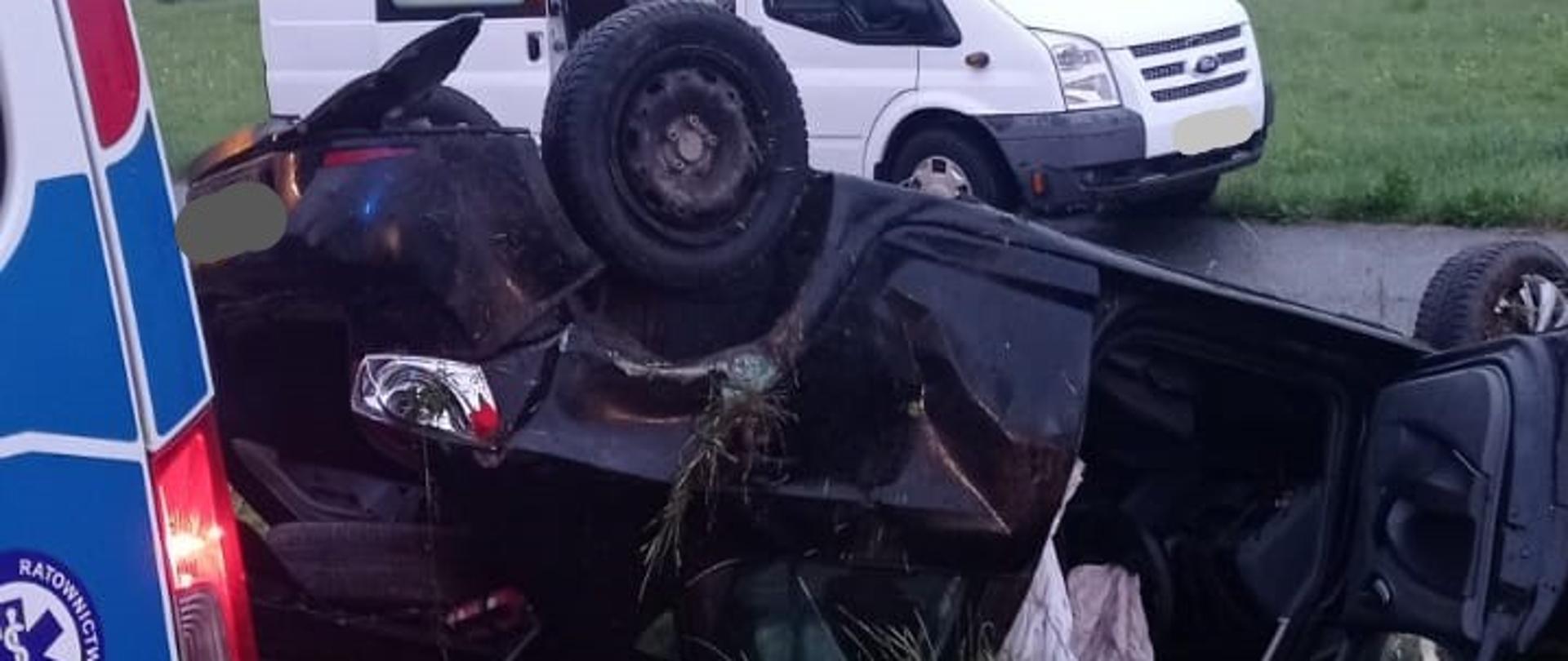 Zdjęcie przedstawia w centralnej części zniszczony samochód osobowy leżący na dachu na nasypie drogi. Z lewej strony widoczny jest tylny narożnik karetki pogotowia. Za zniszczonym pojazdem na drodze lokalnej stoi samochód dostawczy w kolorze białym.