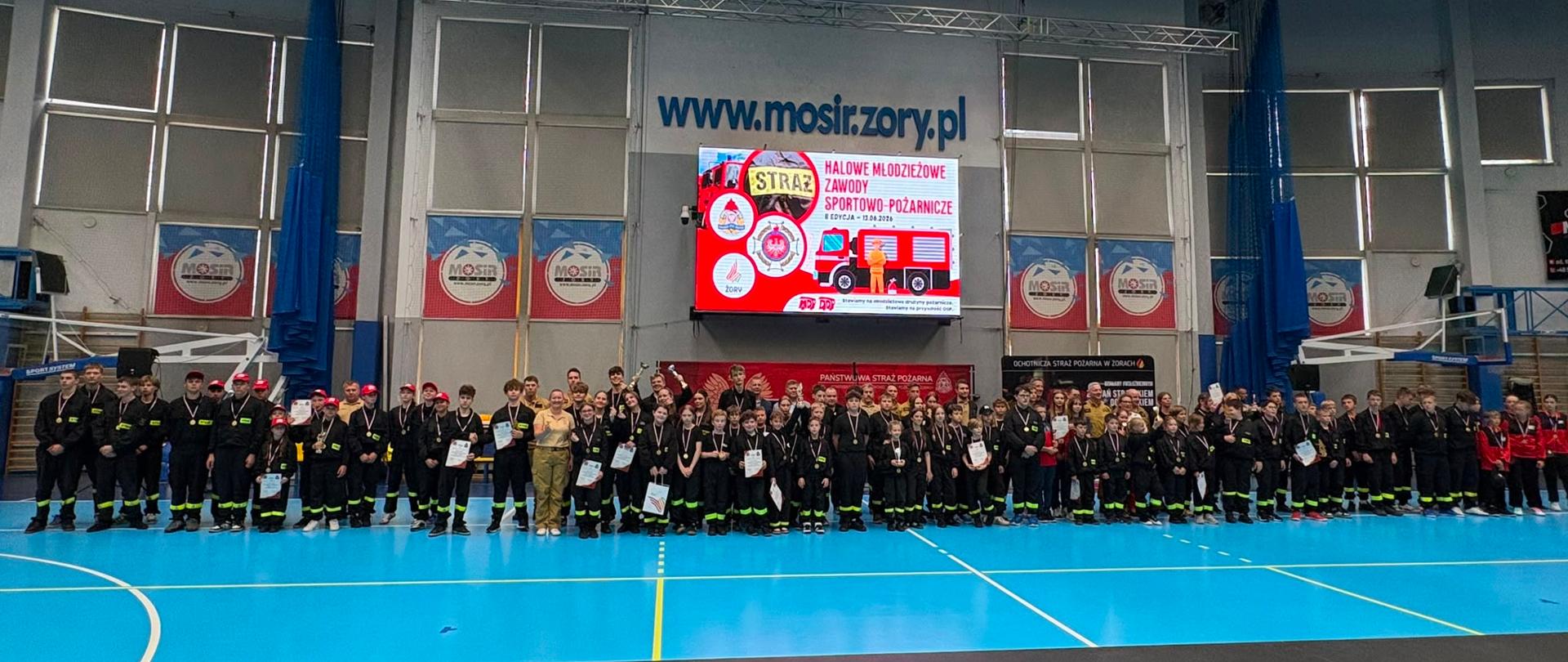 
Uczestnicy II Halowych Młodzieżowych Zawodów Sportowo-Pożarniczych pozują do wspólnego zdjęcia na hali sportowej MOSiR w Żorach, trzymając dyplomy i medale.
