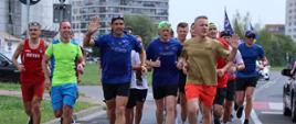 Uczestnicy ultramaratonu biegnący ulicami warszawy. Siedemnastoosobowa grupa ukraińskich i polskich strażaków w strojach sportowych biegnąca ulicą. Na pierwszym planie komendant główny i ukraiński ratownik inicjator maratonu pozdrawiają osoby postronne. W tle pozostali użytkownicy.