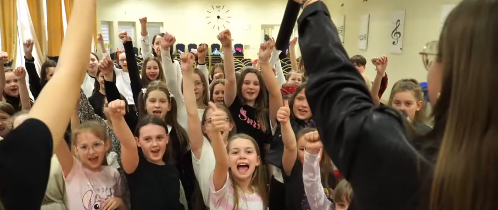 Zbiorowe zdjęcie dzieci z podniesionymi prawymi rękoma do góry z zaciśniętymi piąstkami. Dzieci śpiewają piosenkę o szkole.