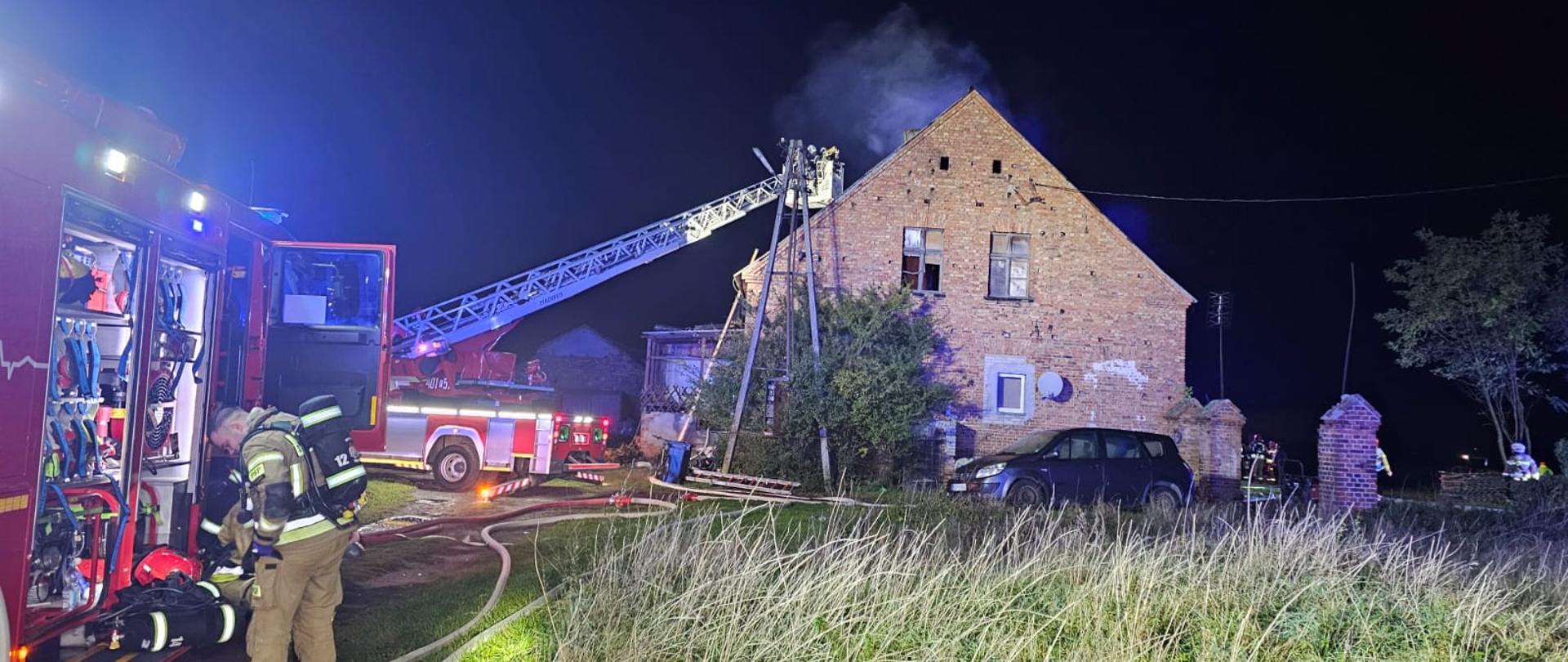 Zdjęcie przedstawia strażaków podczas gaszenia pożaru budynku wielorodzinnego.
W tle samochody strażackie i budynki.
