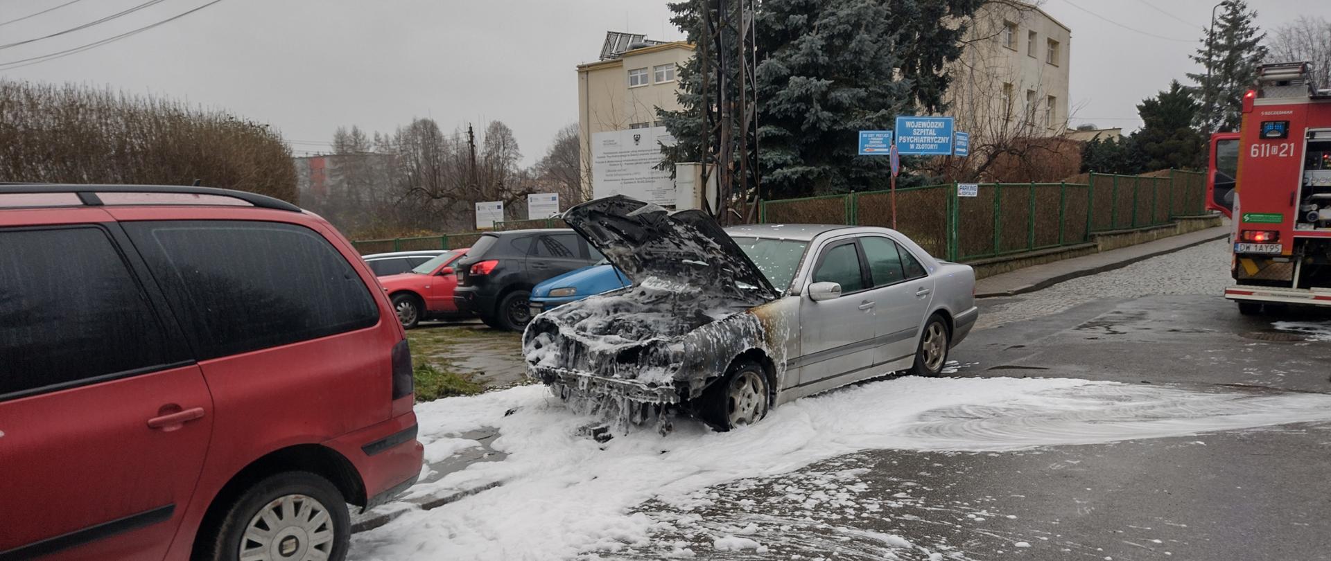 stojący na ulicy spalony do połowy srebrny samochód osobowy zalany pianą gaśniczą, po prawe stronie widoczny strażacki samochód