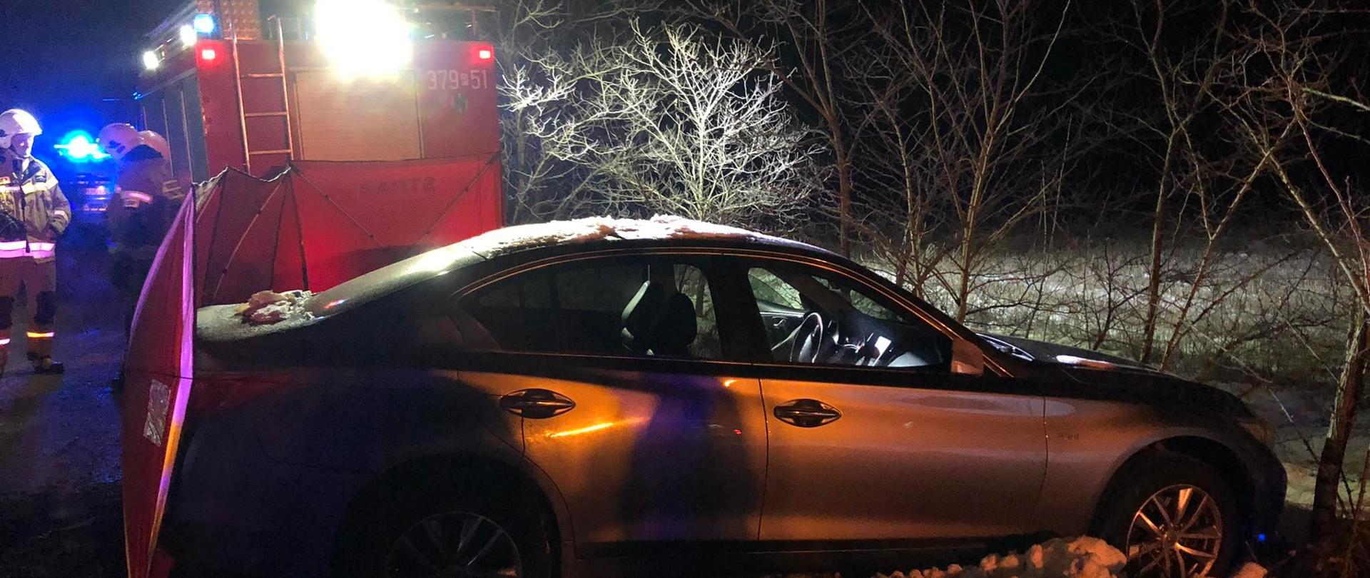 Zdjęcie przedstawia samochód osobowy stojący częściowo na jezdni oraz częściowo w przydrożnym rowie. Pora nocna. Za samochodem rozłożony jest parawan strażacki. Na zdjęciu widać samochód strażacki oraz strażaków. 