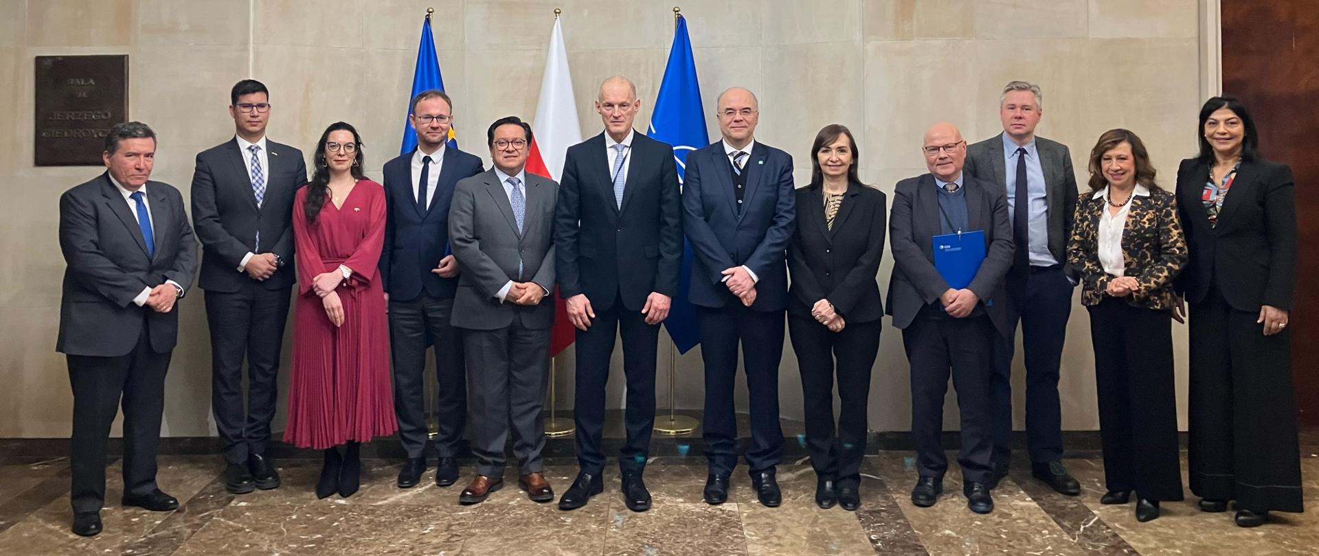 A group of formally dressed individuals pose for a group photo in a representative hall. Flags and a marble wall are visible in the background, with an ornate chandelier above them. The participants stand in a single row, facing the camera.