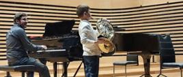 Chłopczyk stojąc na etsradzie sali koncertowej gra na waltorni, za nim na fortepianie Fazioli gra mężczyzna.