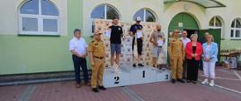 Fotografia przedstawia podium zwycięzców w kategorii mężczyzn wyścigu rowerowego MTB o puchar komendanta powiatowego. Po prawej i lewej stronie widać strażaków Państwowej Straży Pożarnej w ubraniu służbowym stojących obok podium. Wygrani prezentują dyplomy oraz statuetki.