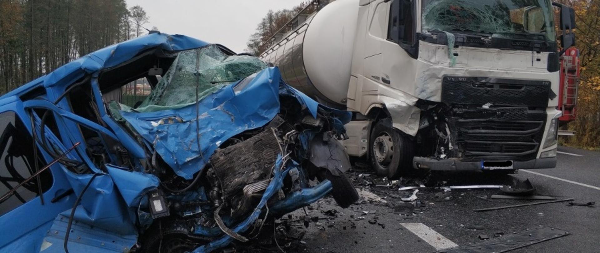 Na zdjęciu widoczne od lewej strony: rozbity bus koloru niebieskiego, częściowo w przydrożnym rowie, przód wystaje na jezdnię; ciągnik siodłowy koloru białego z uszkodzoną przednią szybą oraz uszkodzeniami prawej strony kabiny.