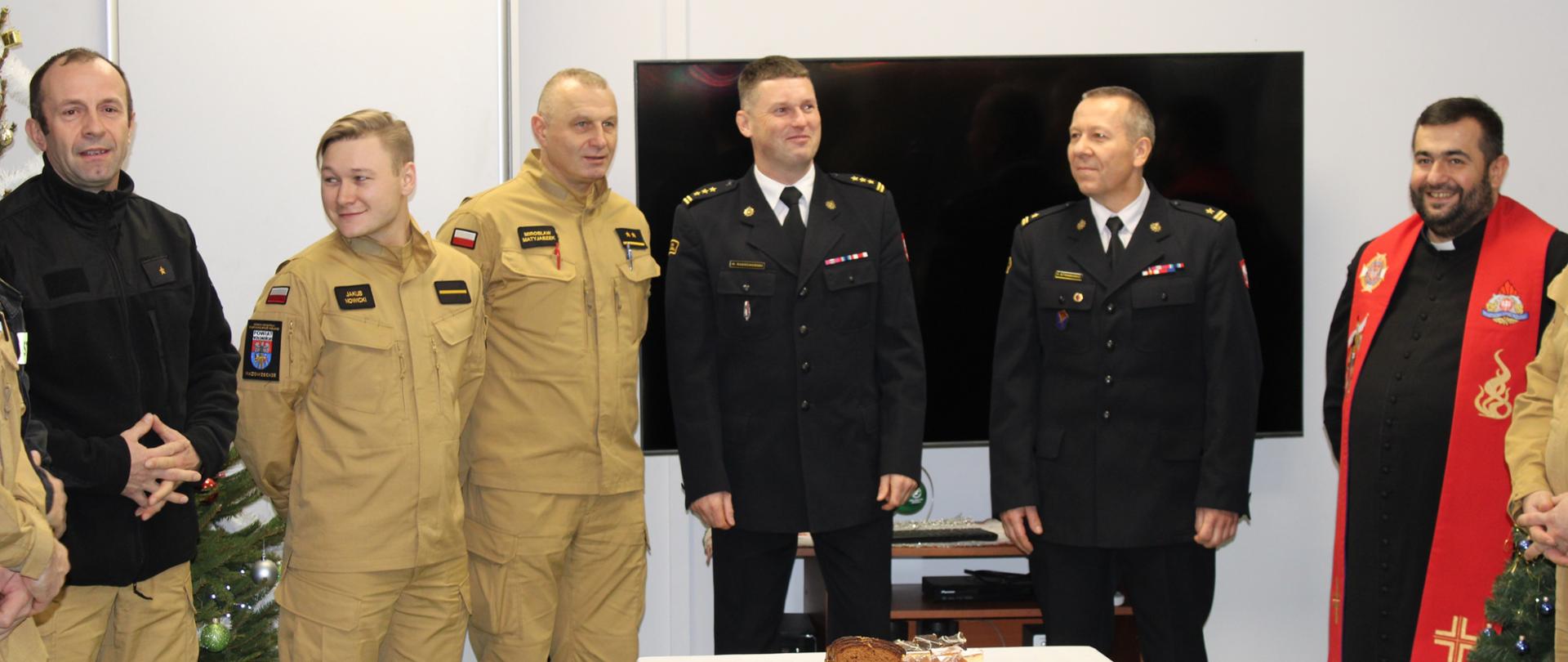 Zdjęcia przedstawiają spotkanie opłatkowe wołomińskich strażaków. Spotkanie odbywa się na świetlicy JRG. Na zdjęciach widać Komendantów oraz księdza kapelana. Strażacy ubrani są w mundury służbowe, koszarowe i wyjściowe.