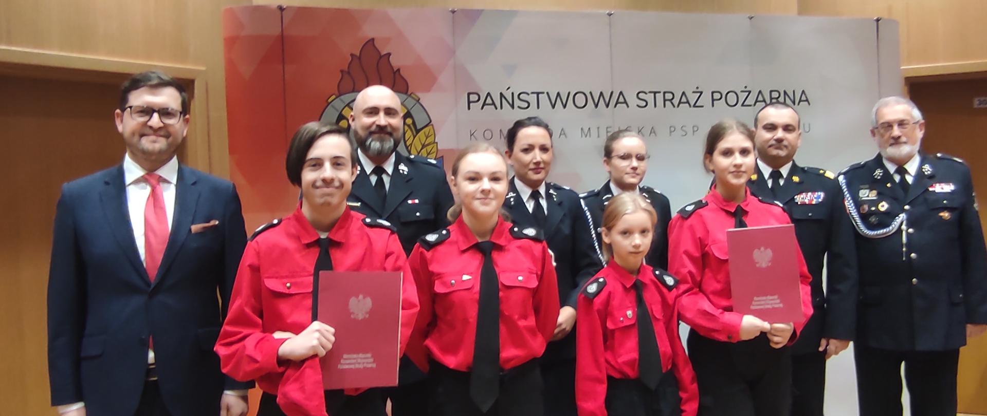 Zdjęcie zrobione w pomieszczeniu, na tle baneru Państwowa Straż Pożarna. Do zdjęcia pozują z lewej przedstawiciel rządu, dalej strażacy w mundurach galowych oraz w czerwonych mundurach czwórka dzieci z młodzieżowych drużyn pożarniczych.