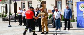Komendant Miejski ubrany w piaskowy mundur wręcza statuetkę uczestnikowi zawodów ubranemu w czerwony podkoszulek z napisem straz i czarne spodnie. Z tyłu stoi kilkanaście osób w tle budynki w zabudowie szeregowej