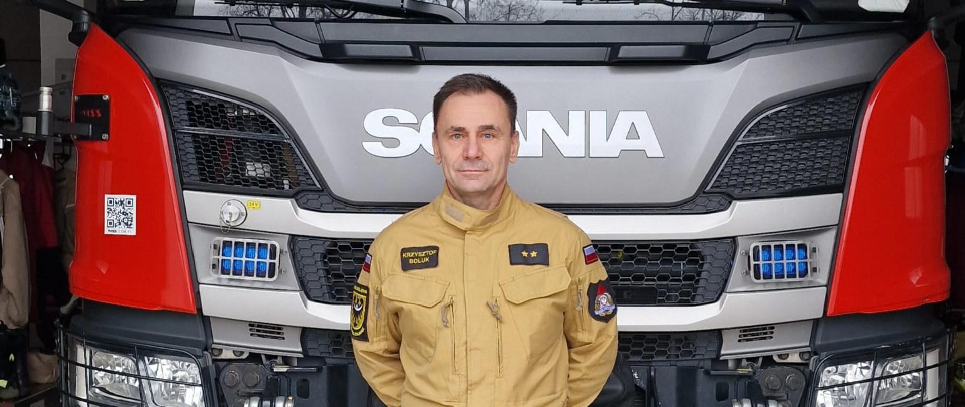 Zdjęcie przedstawia strażaka w piaskowym (beżowym) mundurze służbowym, stojącego na pierwszym planie, dokładnie na środku kadru. Pozuje on na tle przodu nowoczesnego, dużego wozu strażackiego marki Scania zaparkowanego w garażu jednostki ratowniczo-gaśniczej.