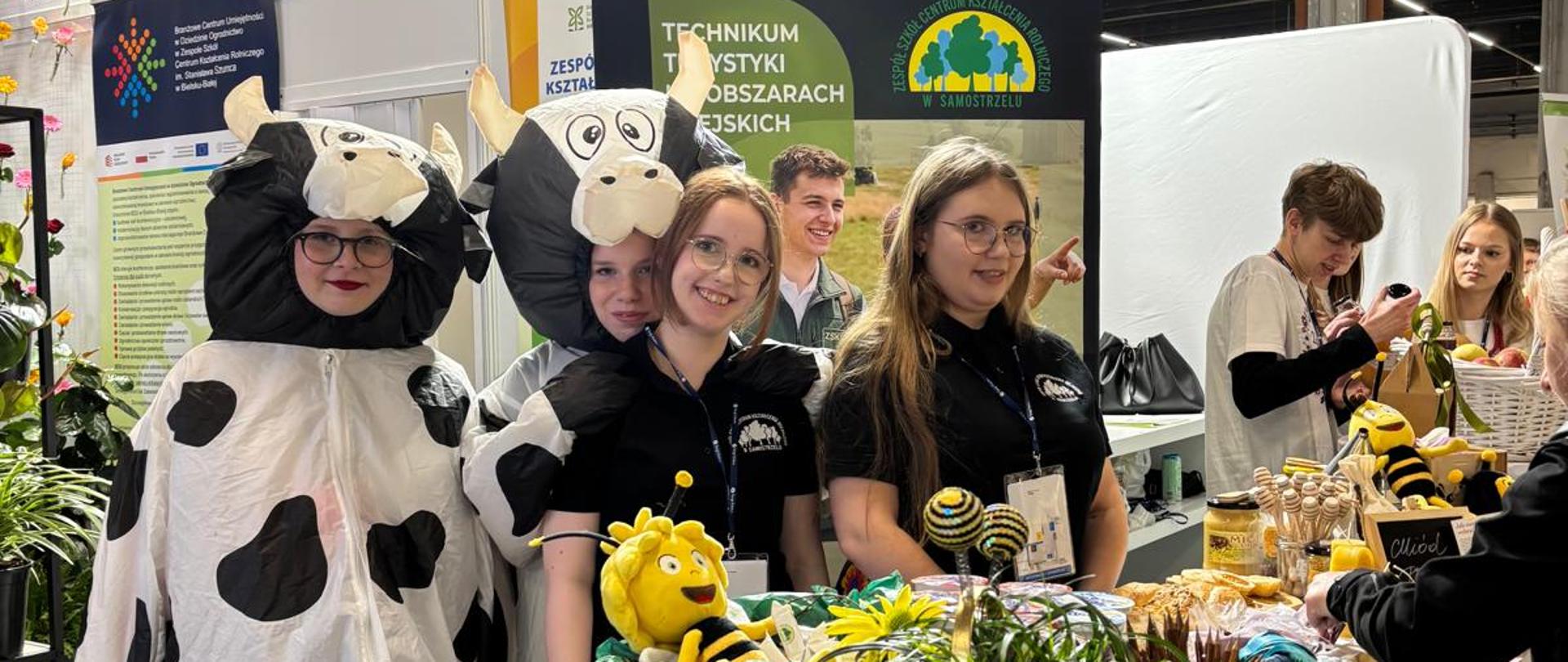 Uczennice ZSCKR w Samostrzelu na stoisku targowym Agro&Activ w Kielcach. Dwie osoby ubrane są w zabawne stroje krów, a pozostałe uczennice uśmiechają się do obiektywu w czarnych koszulkach z logo szkoły. Na pierwszym planie biały stół z dekoracjami: koszem z maskotką pszczoły, miodem i kwiatami. W tle widoczne banery Technikum Turystyki na Obszarach Wiejskich oraz Sieci Szkół Rolniczych.