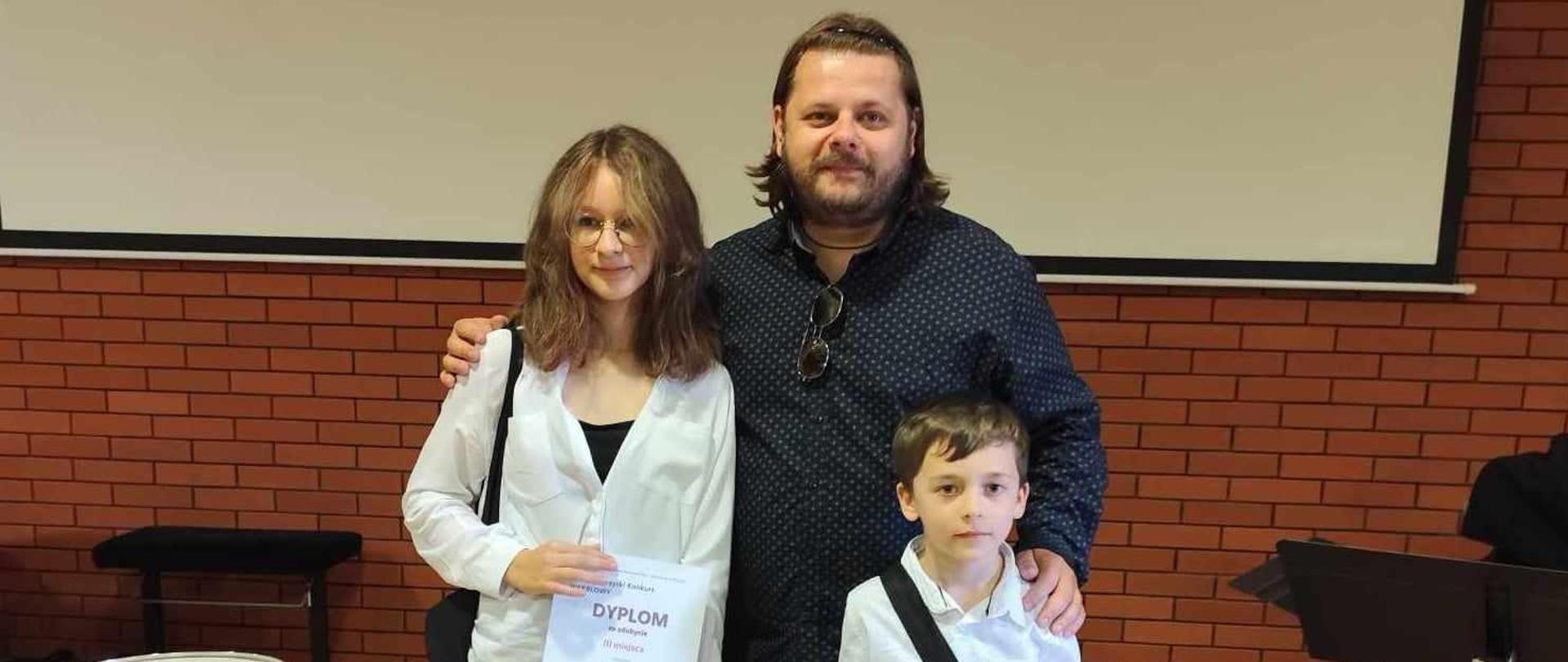 Na środku zdjęcia widać nauczyciela z uczennicą i uczniem którzy trzymają w rękach dyplomy w tle widać tablicę oraz ścianę z cegieł. Po lewej stronie na zdjęciu widać również werbel a po prawej stronie kilka pulpitów oraz krzesło od fortepianu