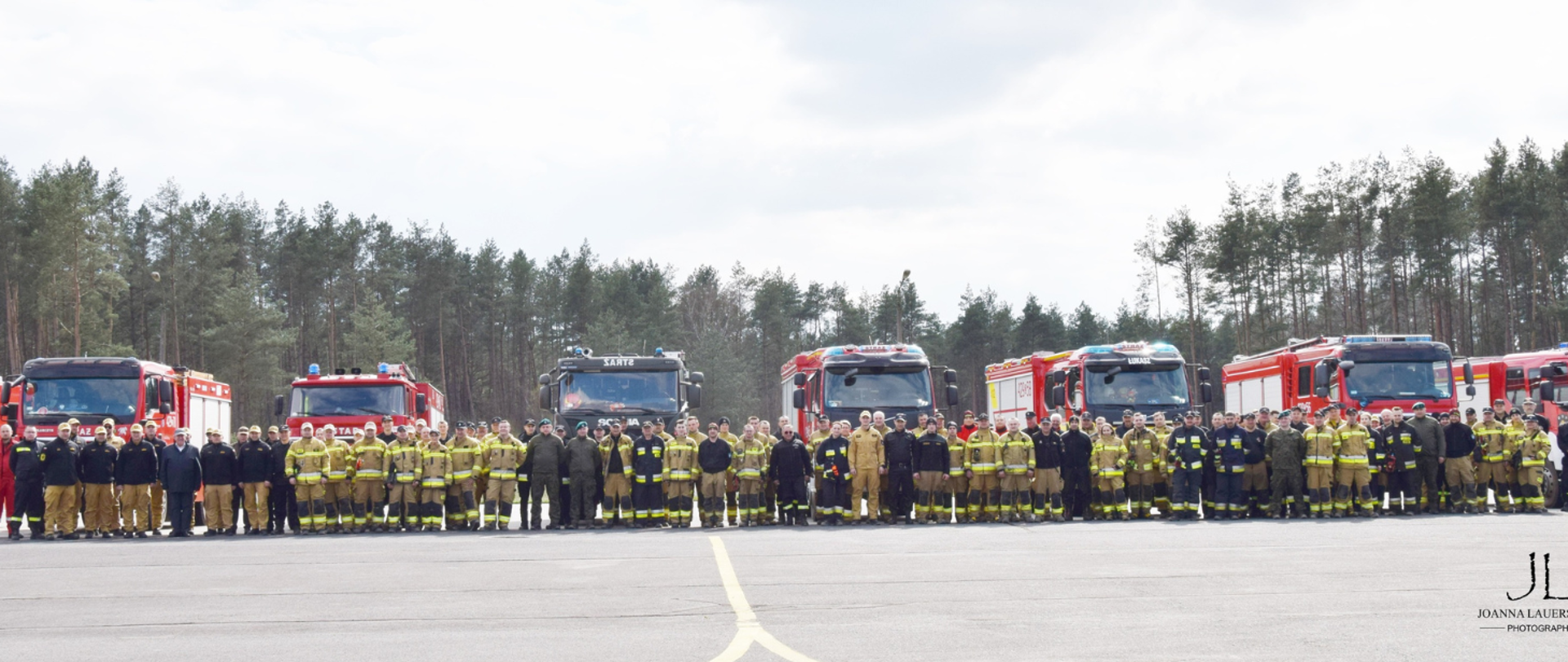 Widok panoramiczny strażaków biorących udział w manewrach na tle samochodów pożarniczych i lasu 