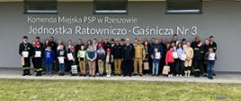 Zdjęcie przedstawia uczestników konkursu trzymających dyplomy, medale oraz puchary, opiekunów oraz organizatorów konkursu podczas pamiątkowej fotografii na zakończenie ogólnopolskiego konkursu wiedzy pożarniczej. Zdjęcie zrobione jest przed budynkiem Jednostki Ratowniczo-Gaśniczej nr 3 w Rzeszowie.