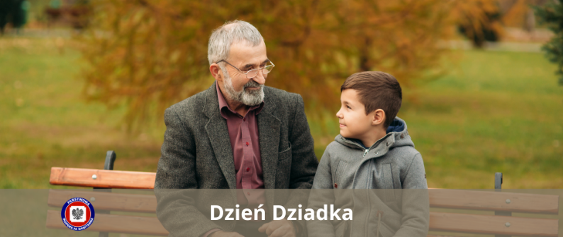 Na ławce w parku siedzi dziadek z wnuczkiem, patrzą na siebie uśmiechnięci. 