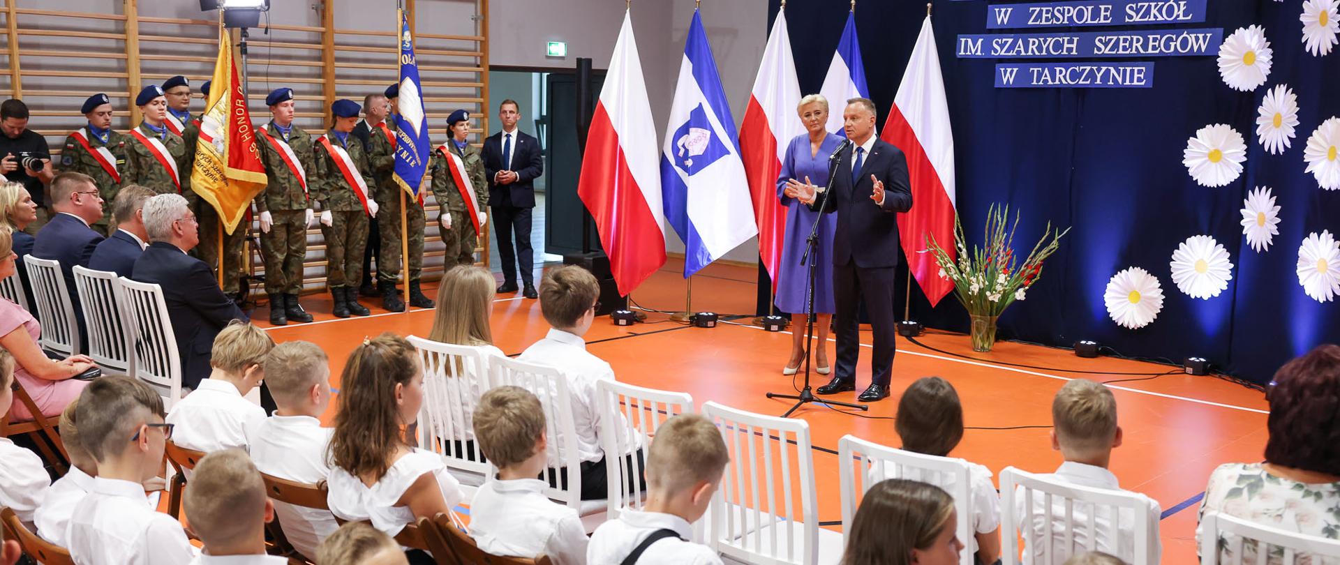 Przy mikrofonie para prezydencka, po lewej stronie uczniowie ubrani w mundury, na pierwszym planie dzieci ubrane w białe bluzki i koszule 