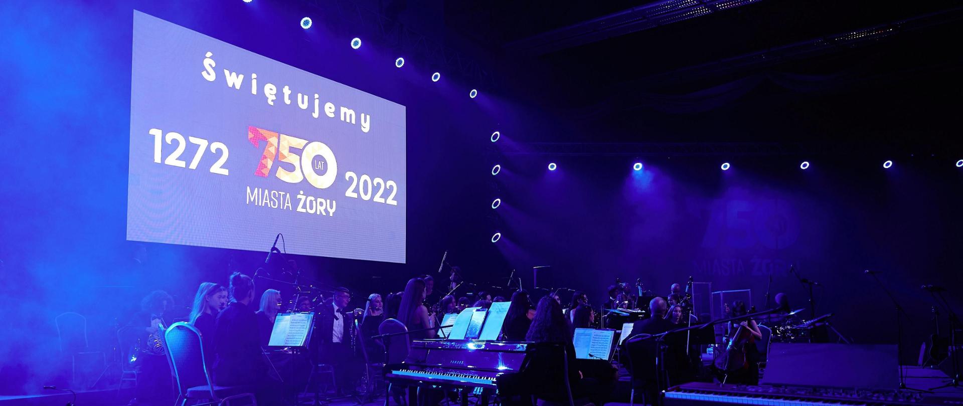 W lewym górnym rogu duży telebim z napisem świętujemy 750 lat miasta Żory 1272 do 2022, poniżej na scenie młodzieżowa orkiestra symfoniczna w trakcie koncertu w ramach obchodów rocznicowych.