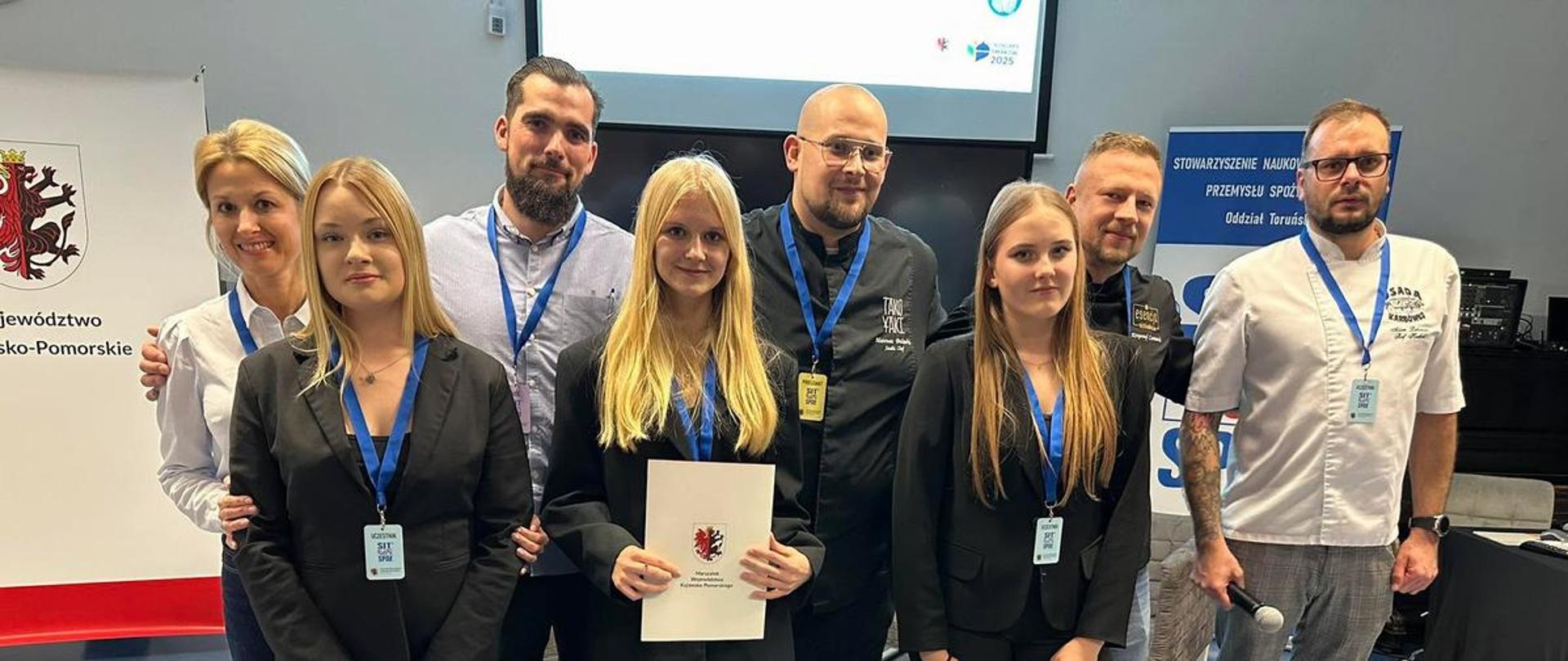 Zdjęcie przedstawia grupę siedmiu osób, pozujących do wspólnego zdjęcia wewnątrz budynku, prawdopodobnie podczas wydarzenia branżowego lub edukacyjnego.
Pierwszy plan: W rzędzie z przodu stoją trzy młode kobiety, wszystkie ubrane w jednakowe czarne marynarki i spodnie. Środkowa kobieta trzyma w rękach biały dokument lub certyfikat z herbem. Wszyscy w grupie noszą zawieszone na szyi smycze z identyfikatorami.
Tło: Za kobietami stoją dorośli – dwie kobiety i trzech mężczyzn. Wśród nich są osoby ubrane w koszule, marynarki, a po prawej stronie widać dwóch mężczyzn w profesjonalnych kitlach szefa kuchni.
Otoczenie: W tle widoczne są elementy wystroju, w tym ekran projekcyjny i fragmenty tablic informacyjnych. Na tablicy po lewej widoczny jest herb województwa kujawsko-pomorskiego, a na innej tablicy po prawej tekst: "STOWARZYSZENIE NAUKOWE PRZEMYSŁU SPOŻYWCZEGO Oddział Toruński"