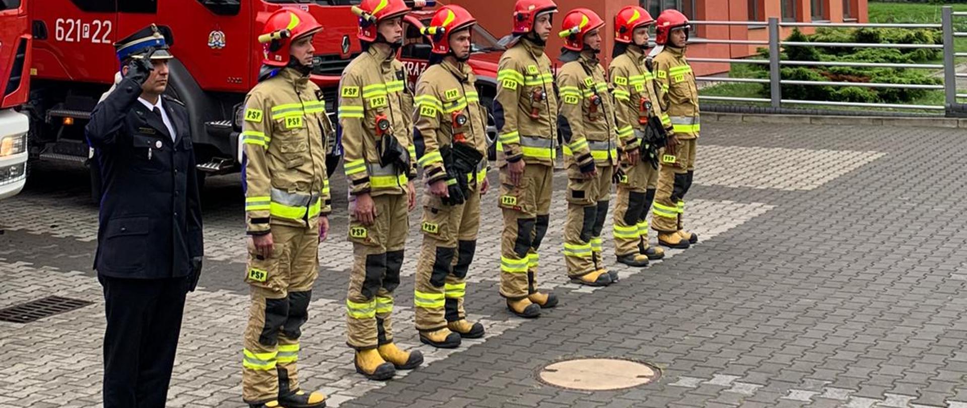 Komendant wraz z zmiana służbową stoją w szeregu przed komendą powiatową w tle samochody pożarnicze.