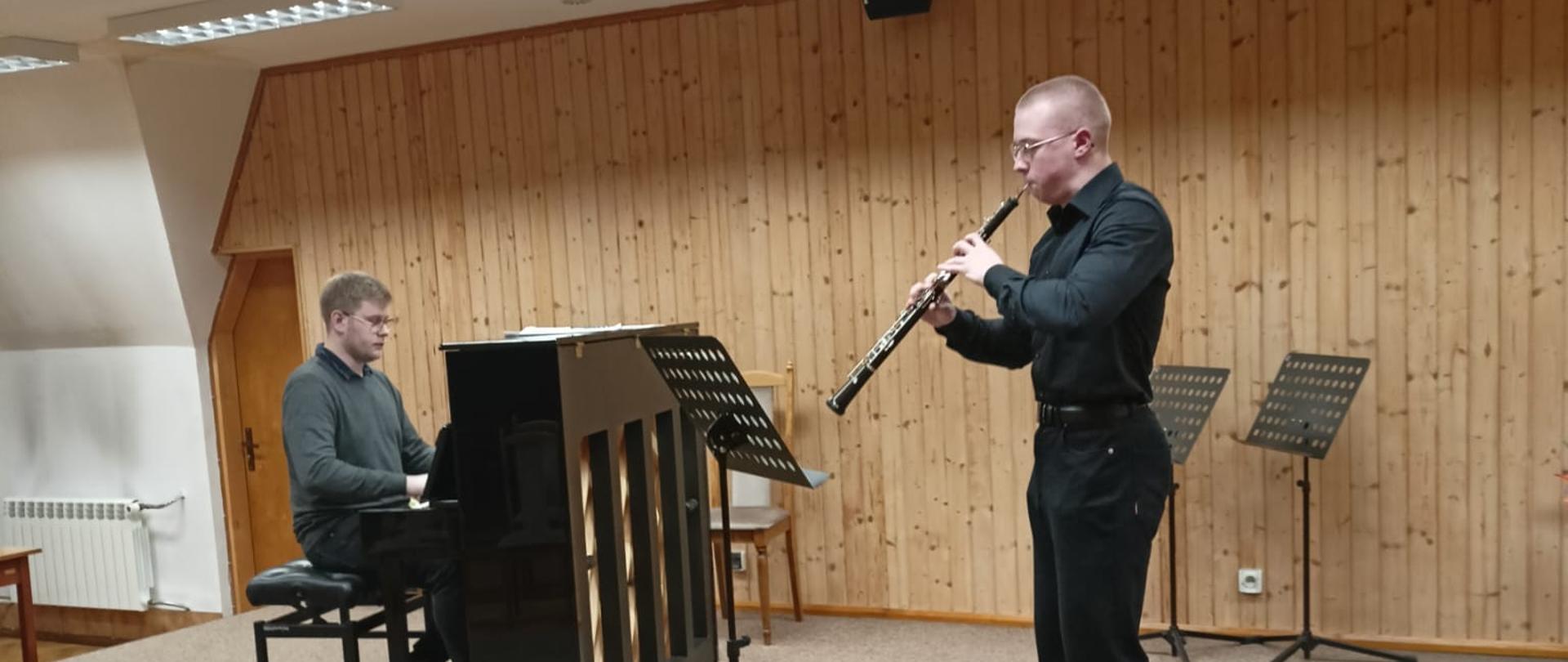 Nastolatek gra na oboju, obok po lewej stronie mężczyzna gra na pianinie.