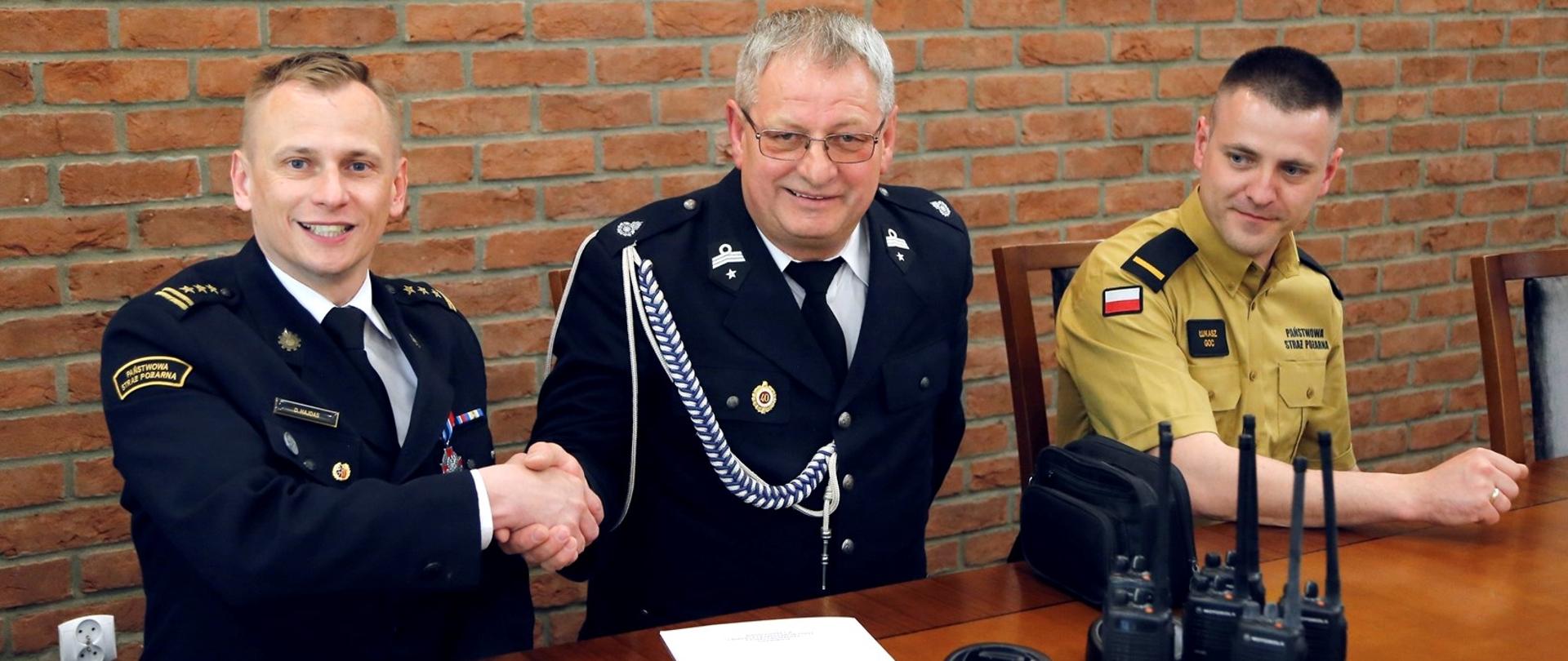Siedzący za stołem Zastępca Komendanta Centralnej Szkoły PSP w uścisku dłoni z siedzącym obok druhem OSP obok siedzący funkcjonariusza wydziału informatyki i łączności CS PSP. Przed nimi na blacie przekazywany sprzęt łączności tj. sześć radiotelefonów nasobnych oraz dwie ładowarki jednostanowiskowe Motorola
